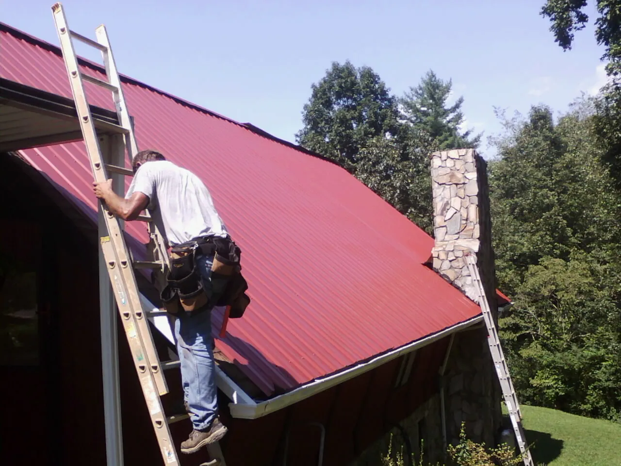 Roofer on a ladder beside a red metal roof and stone chimney — Soffit & Fascia Repair by Roofing Brothers in Ewing